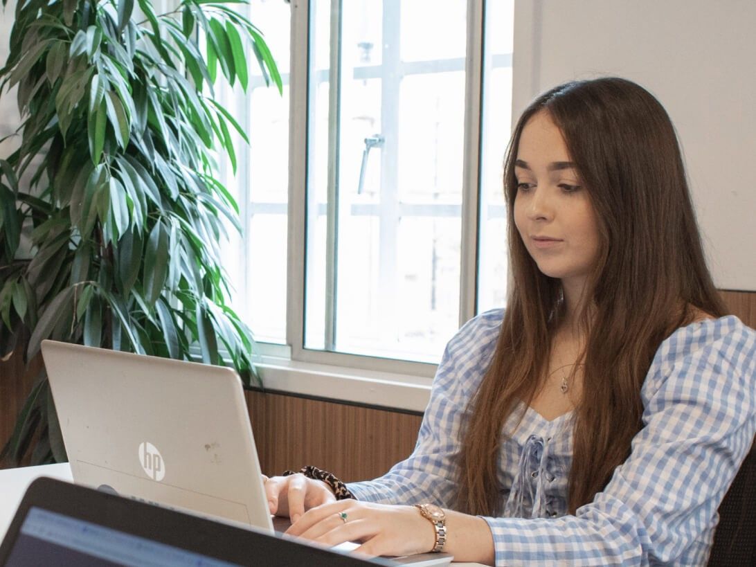 A digital marketing apprentice works on her laptop with windows and plants in the background.